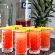Five red-orange cocktails in textured glasses, each garnished with a slice of pineapple, are arranged on a black tray with liquor bottles and a whole pineapple in the background.