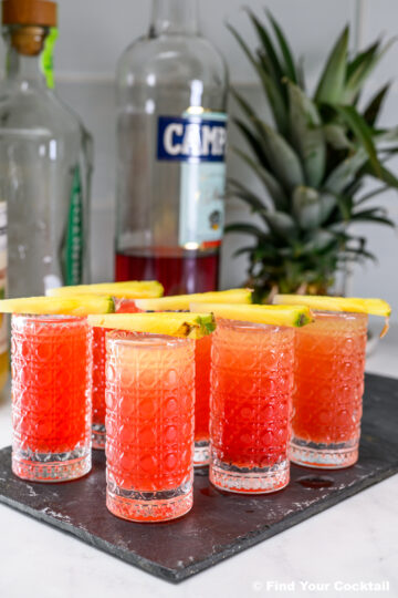 Five red-orange cocktails in textured glasses, each garnished with a slice of pineapple, are arranged on a black tray with liquor bottles and a whole pineapple in the background.