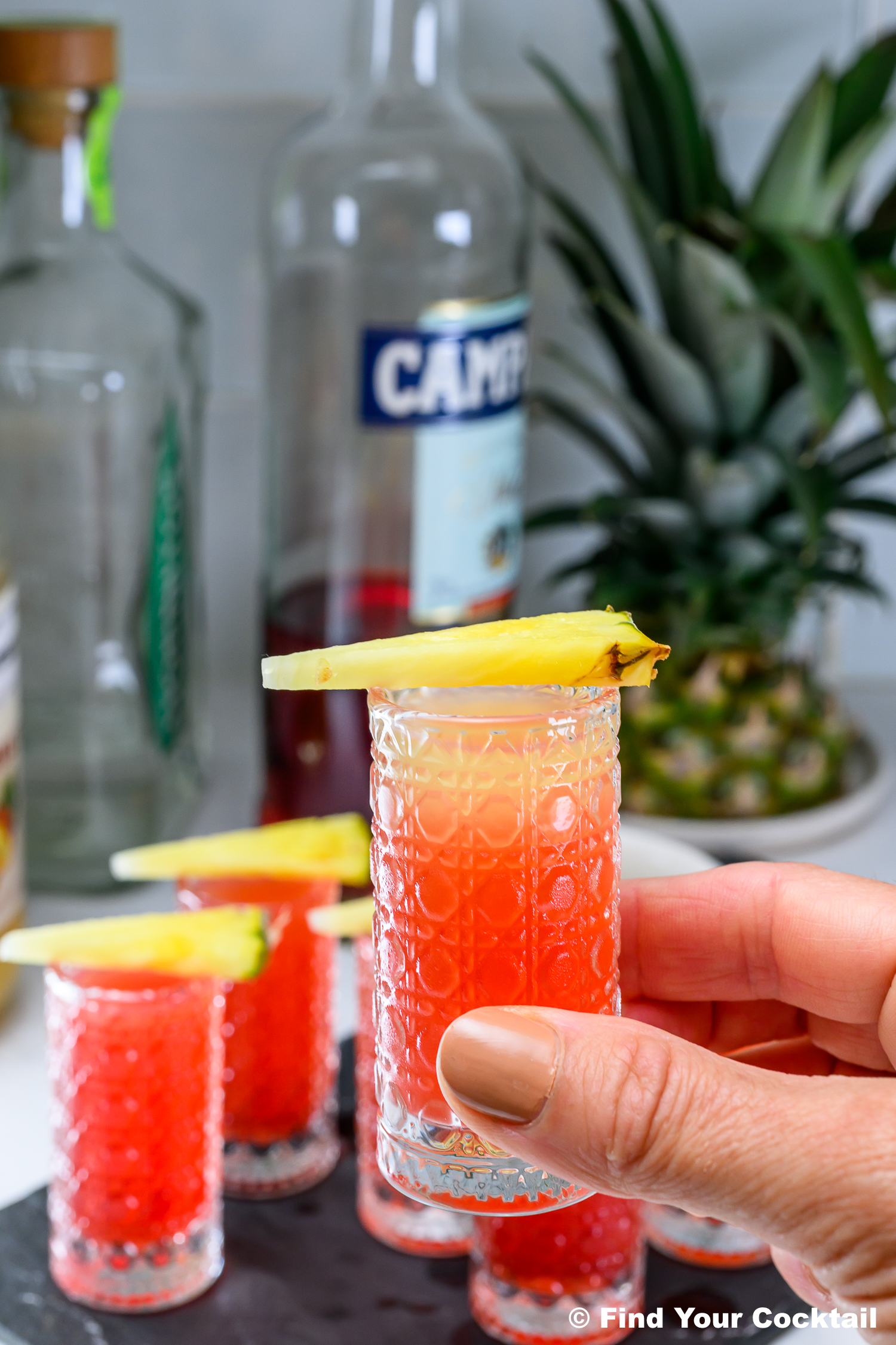 A hand holds a red cocktail in a patterned glass, garnished with a pineapple slice. More drinks, bottles, and a pineapple are visible in the background.