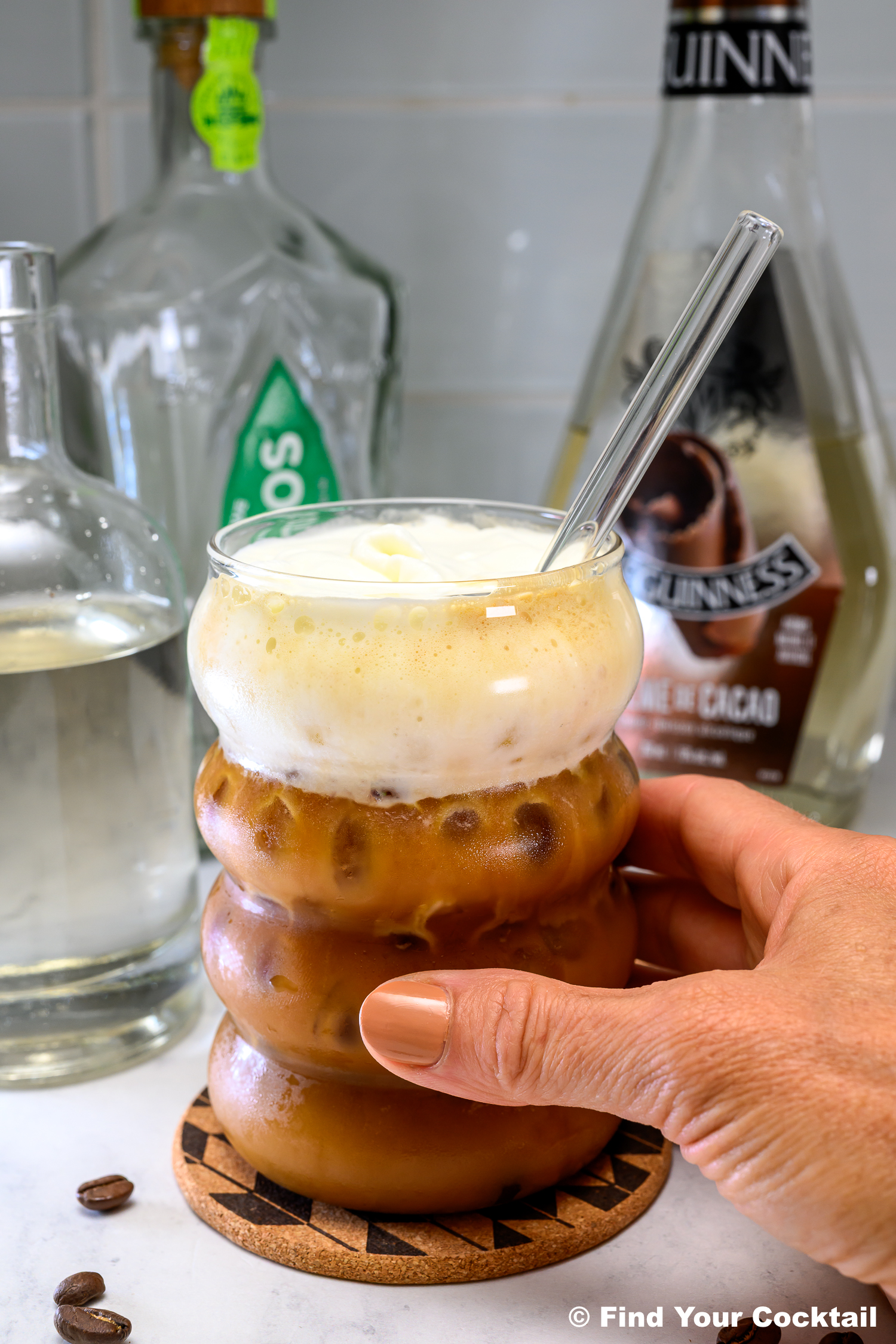 A hand holding a glass of iced coffee topped with foam, with bottles of alcohol and syrup in the background on a countertop.