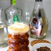 A glass of layered iced coffee with cream on top sits on a coaster, surrounded by coffee beans and bottles of liquor on a countertop.