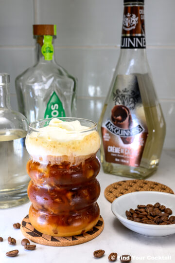 A glass of layered iced coffee with cream on top sits on a coaster, surrounded by coffee beans and bottles of liquor on a countertop.