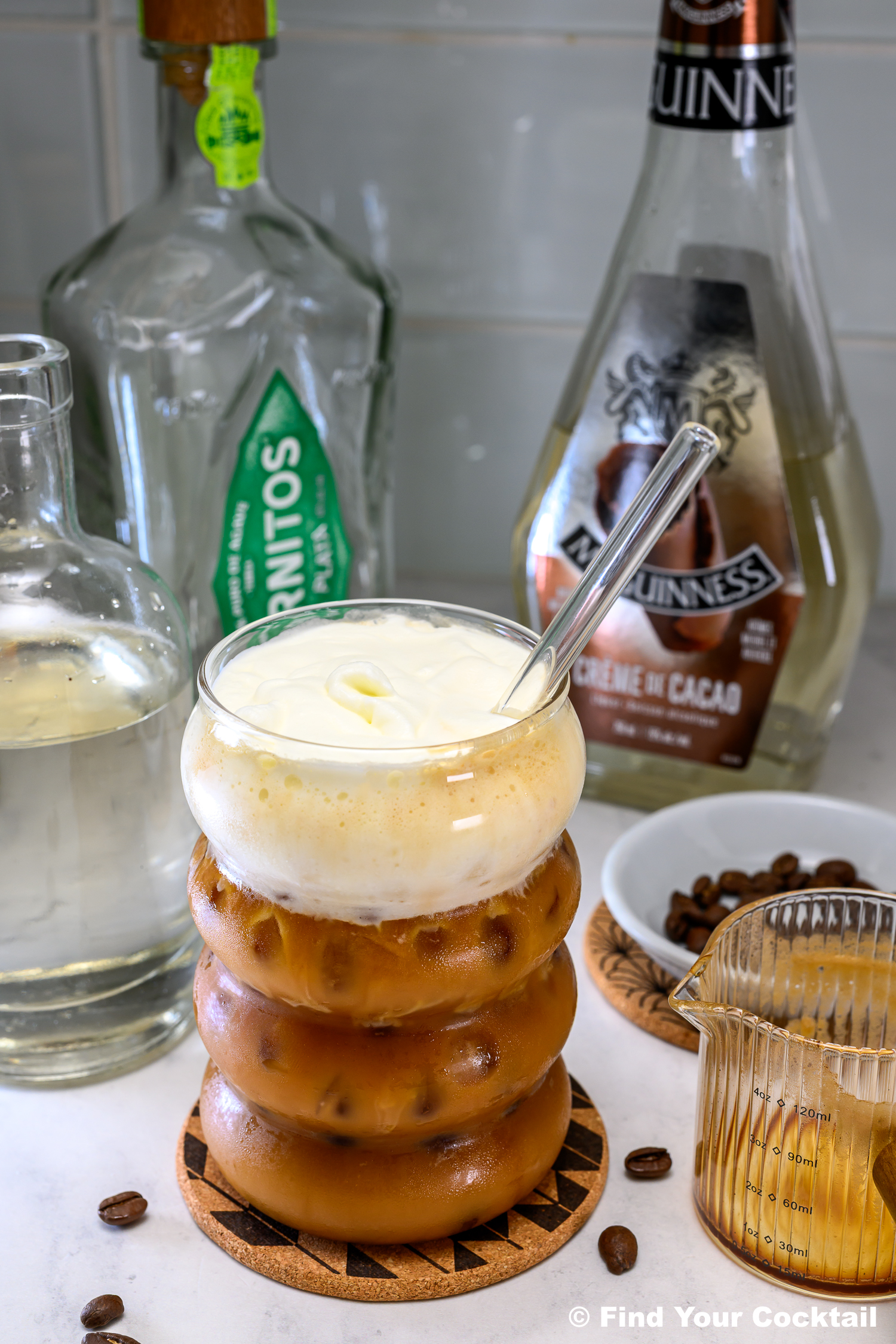 A glass of iced coffee topped with cream sits on a coaster, with bottles of liquor, a glass straw, coffee beans, and a small pitcher nearby on a kitchen counter.