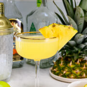 A yellow cocktail garnished with two pineapple slices in a coupe glass, surrounded by fresh pineapple, lime, bottles, a cocktail shaker, and a small bowl.