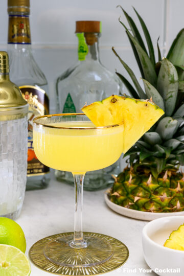 A yellow cocktail garnished with two pineapple slices in a coupe glass, surrounded by fresh pineapple, lime, bottles, a cocktail shaker, and a small bowl.