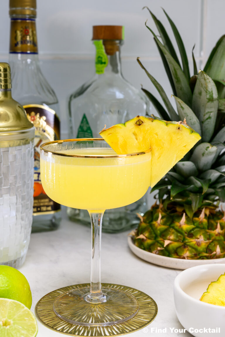 A yellow cocktail garnished with two pineapple slices in a coupe glass, surrounded by fresh pineapple, lime, bottles, a cocktail shaker, and a small bowl.