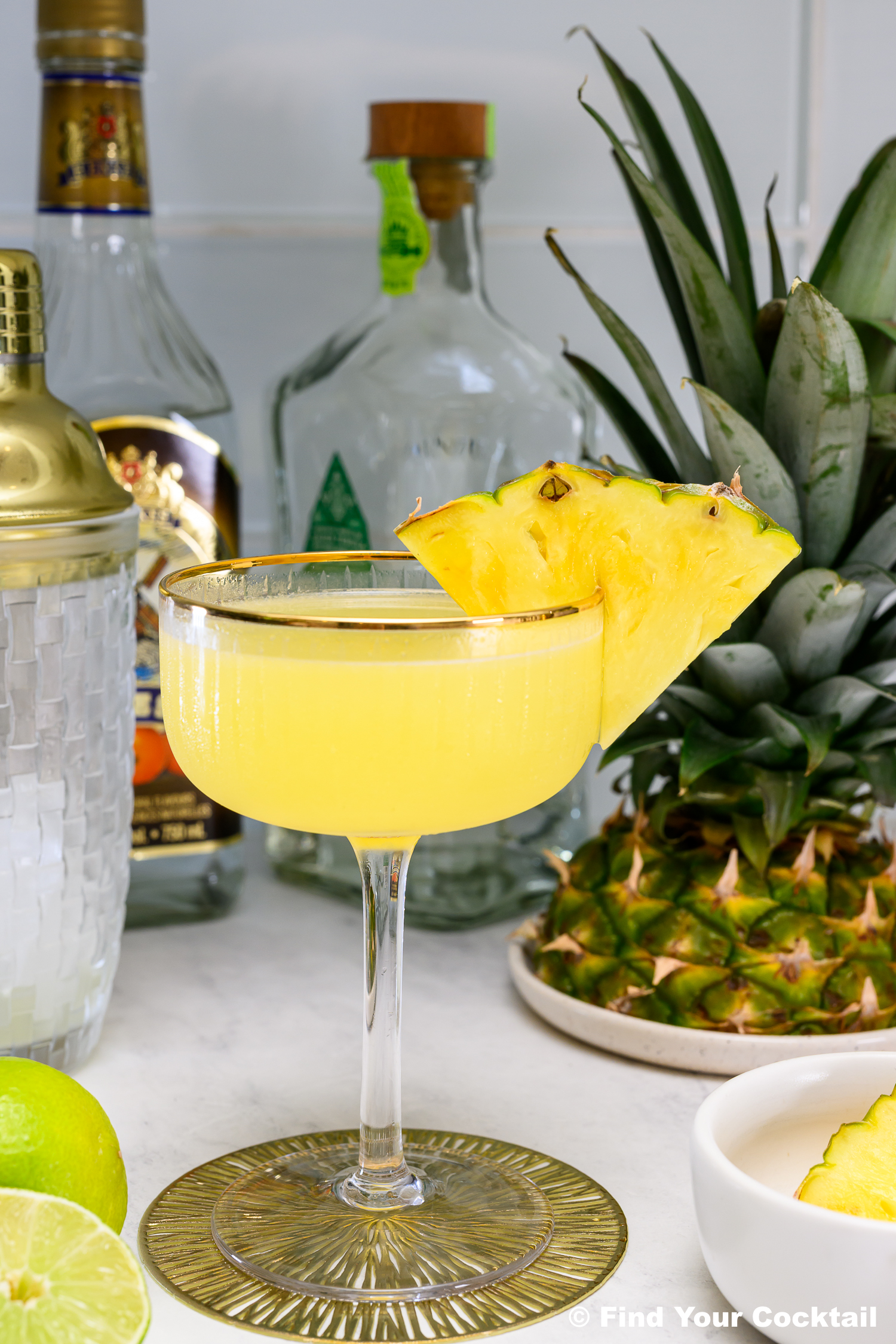 A yellow cocktail garnished with two pineapple slices in a coupe glass, surrounded by fresh pineapple, lime, bottles, a cocktail shaker, and a small bowl.