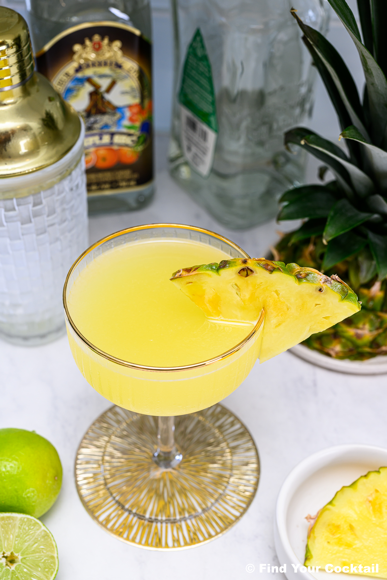 A yellow cocktail garnished with a pineapple slice in a coupe glass, surrounded by bottles, a cocktail shaker, a lime, and a pineapple on a white surface.