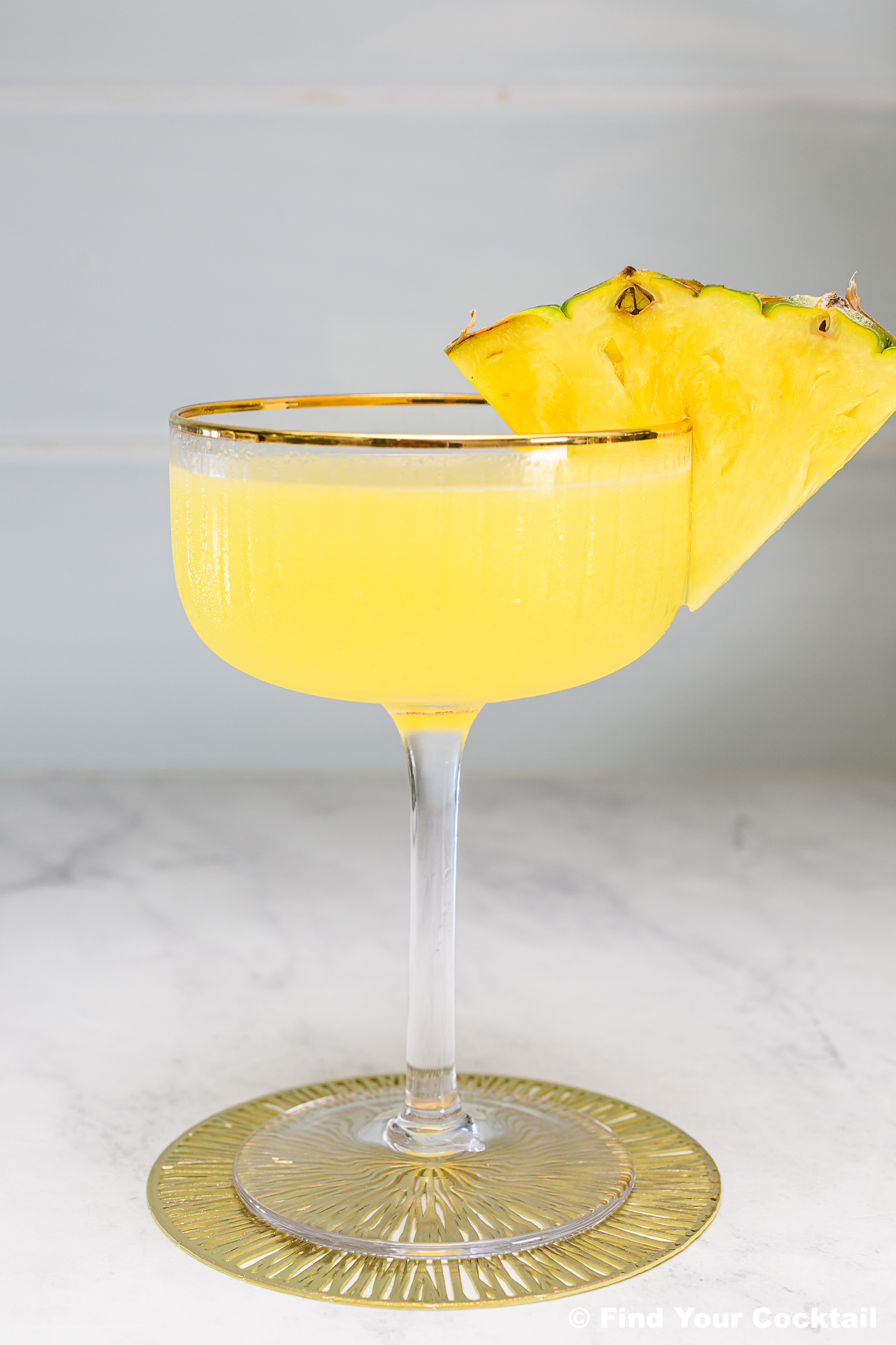 A coupe glass filled with a yellow cocktail, garnished with two pineapple wedges on the rim, sits on a marble surface with a gold coaster underneath.
