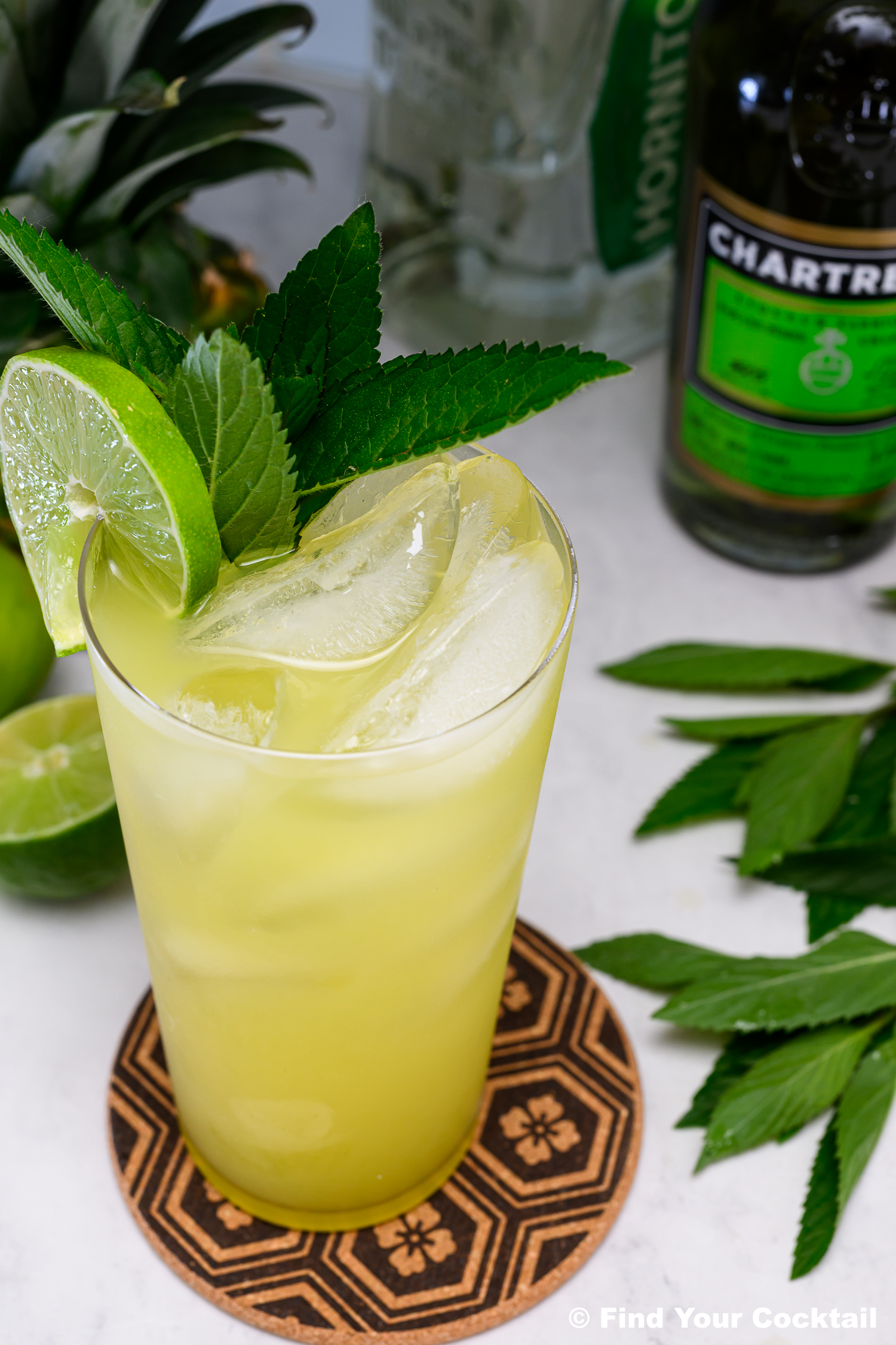 A yellow-green cocktail with ice, garnished with lime slices and mint leaves, sits on a patterned coaster. Bottles and fresh ingredients are in the background.