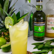 A yellow cocktail with ice, lime wheel, and mint garnish in a tall glass, surrounded by bottles of Chartreuse, tequila, pineapple juice, a lime, and a pineapple.