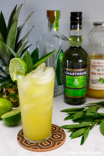 A yellow cocktail with ice, lime wheel, and mint garnish in a tall glass, surrounded by bottles of Chartreuse, tequila, pineapple juice, a lime, and a pineapple.