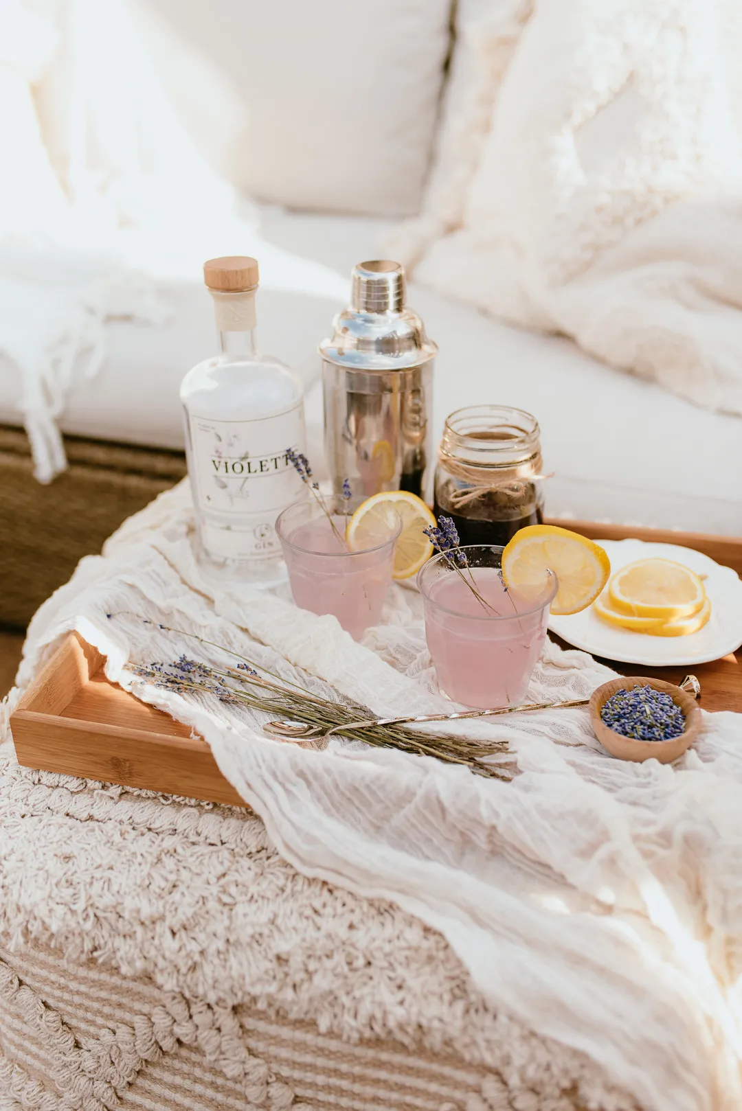 A wooden tray holding two pink cocktails with lemon slices, a bottle of gin, a cocktail shaker, a jar, a plate of lemon slices, and sprigs of lavender on a textured surface.