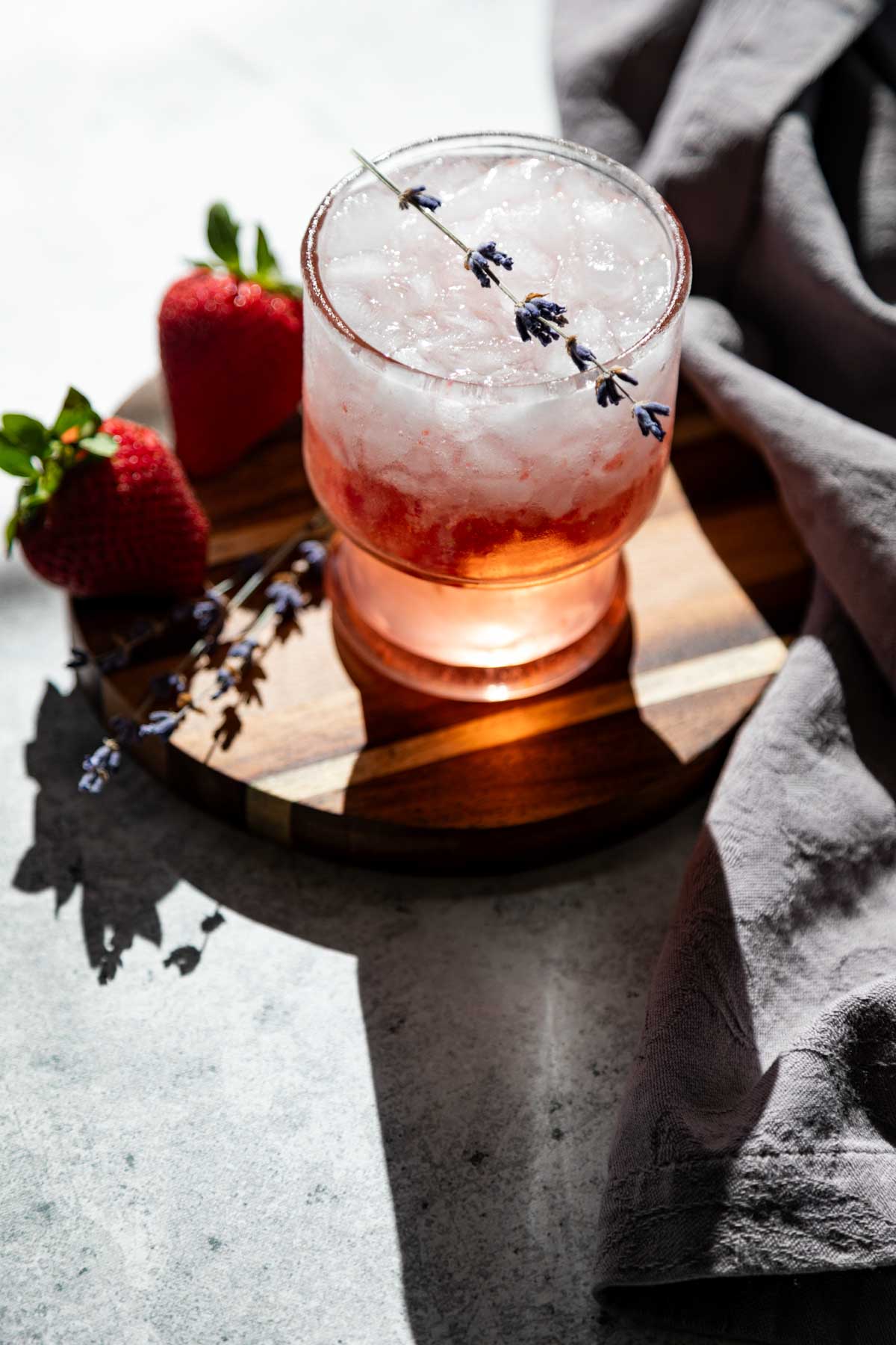 A glass of iced pink drink garnished with a sprig of lavender, placed on a wooden coaster with two strawberries and a gray cloth nearby.