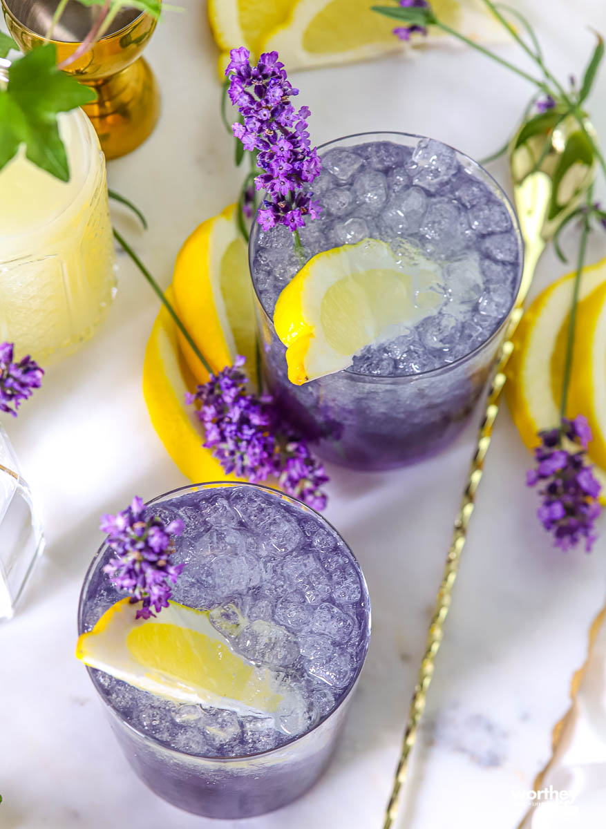 Two glasses of purple iced drink garnished with lemon slices and lavender sprigs, surrounded by lemon wedges and lavender flowers on a white surface.