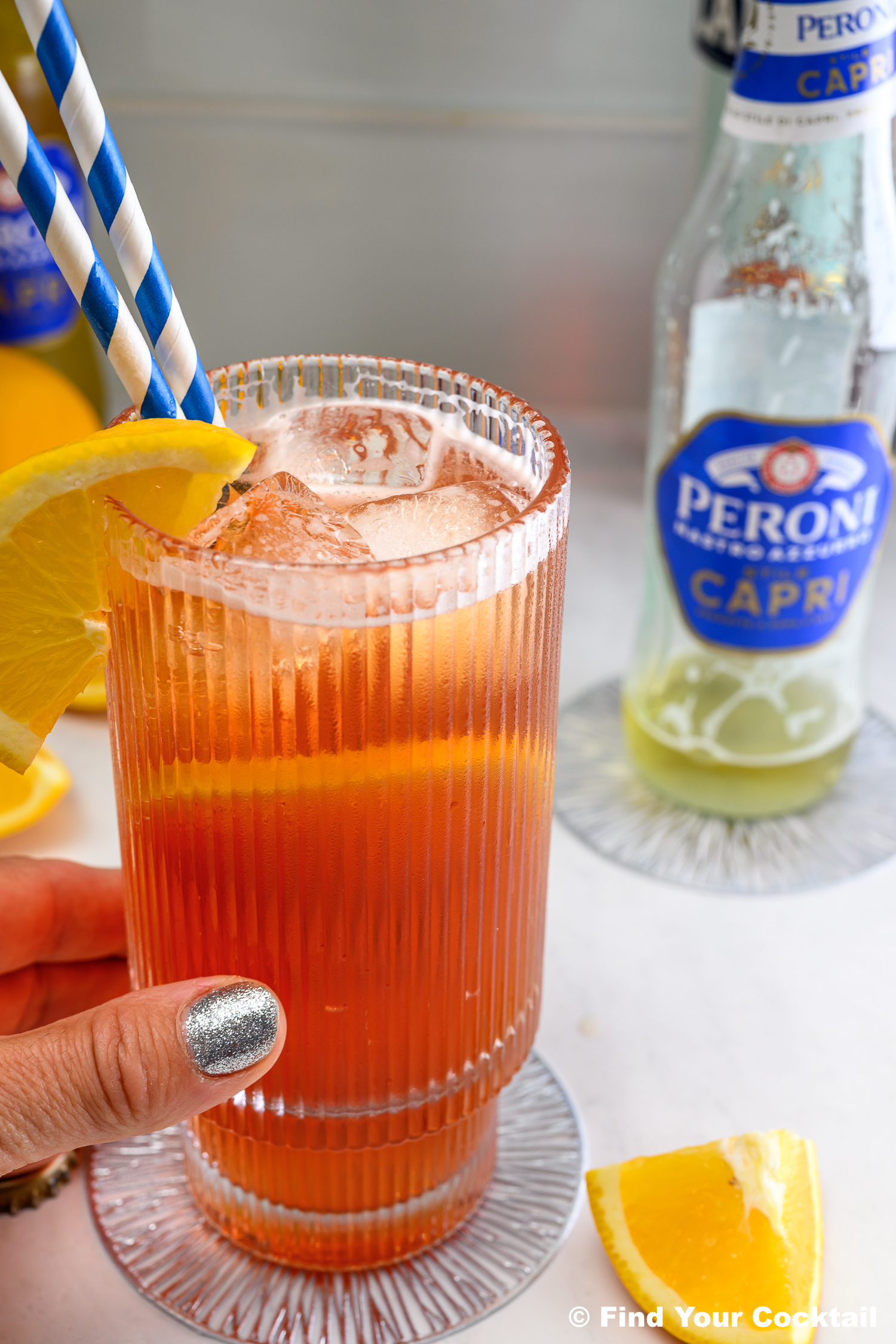 A hand holds a glass of iced orange cocktail with lemon wedge garnish, beside a Peroni Capri bottle, two striped straws, and lemon slices on a white surface.