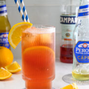 A glass of orange-red cocktail with ice, orange slices, and striped straws sits on a table with bottles, bottle cap, and orange wedges in the background.