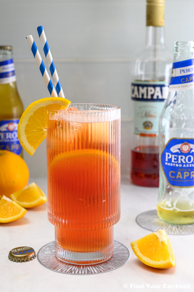 A glass of orange-red cocktail with ice, orange slices, and striped straws sits on a table with bottles, bottle cap, and orange wedges in the background.