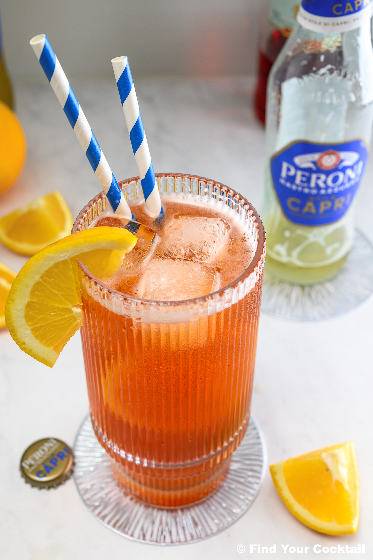 Tall glass of orange cocktail with ice, a lemon slice, and two striped straws; Peroni bottle and orange wedges nearby on a marble surface.