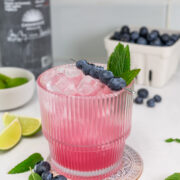 A pink cocktail with ice is garnished with blueberries and mint. Blueberries, lime wedges, and a bottle are in the background on a white surface.
