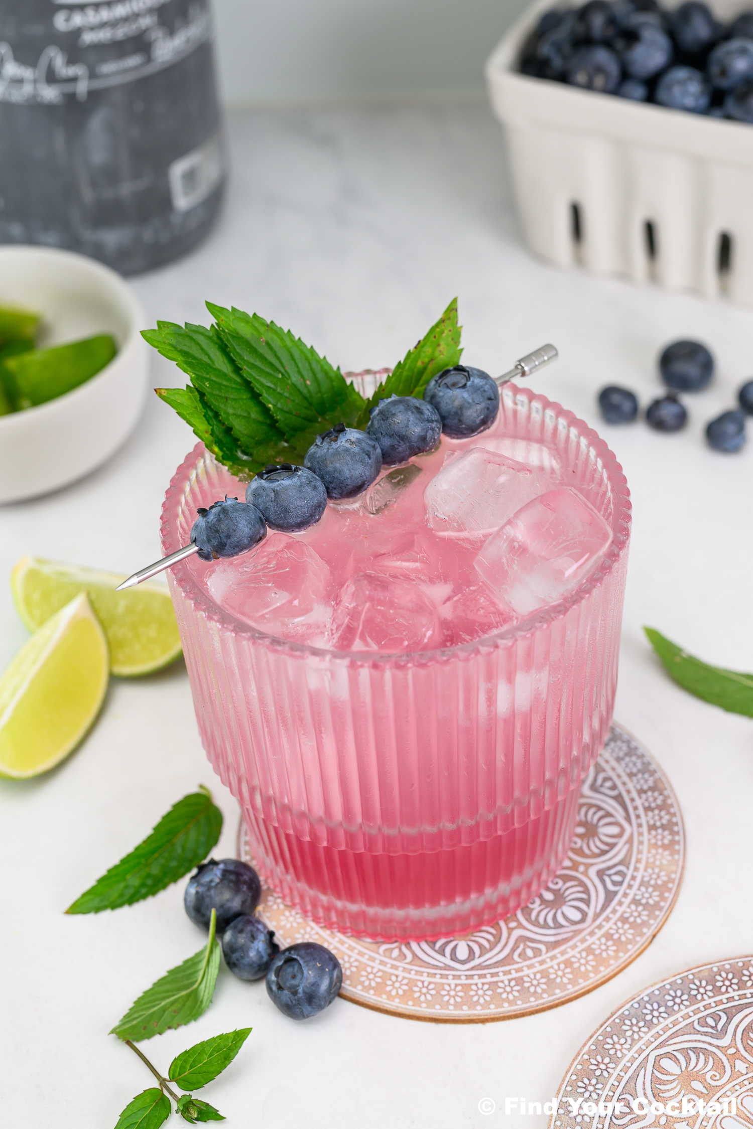A pink cocktail with ice, garnished with mint leaves and blueberries on a skewer, sits on a coaster surrounded by limes and loose blueberries.