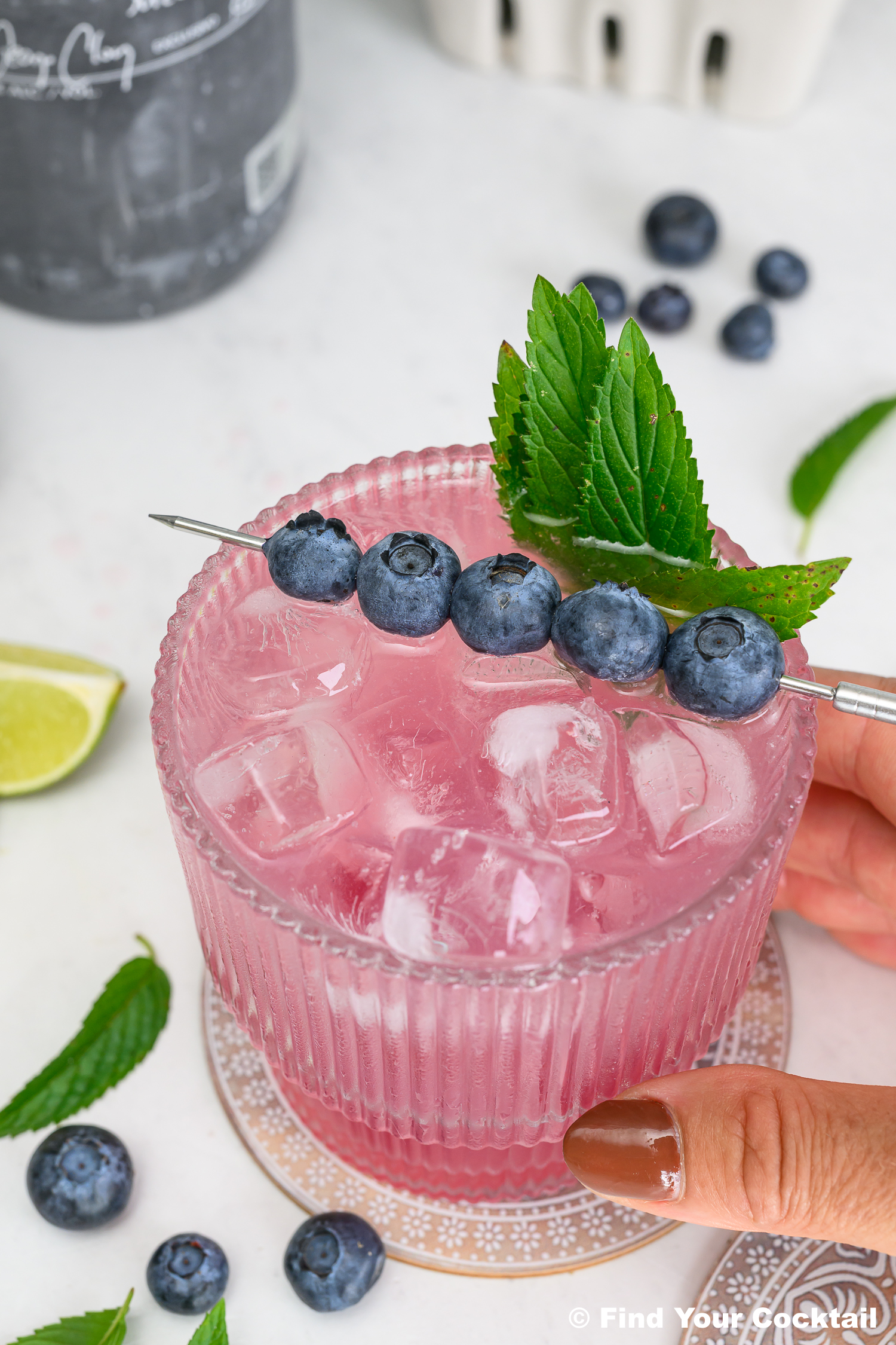 A pink cocktail with ice in a ribbed glass, garnished with blueberries on a skewer and fresh mint, held by a hand with painted nails. Lime, mint, and blueberries are nearby.