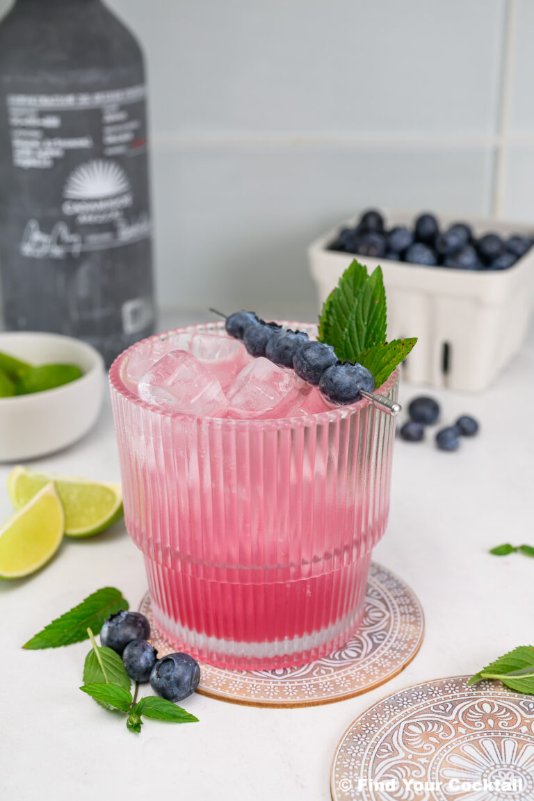 A pink cocktail with ice is garnished with blueberries and mint. Blueberries, lime wedges, and a bottle are in the background on a white surface.