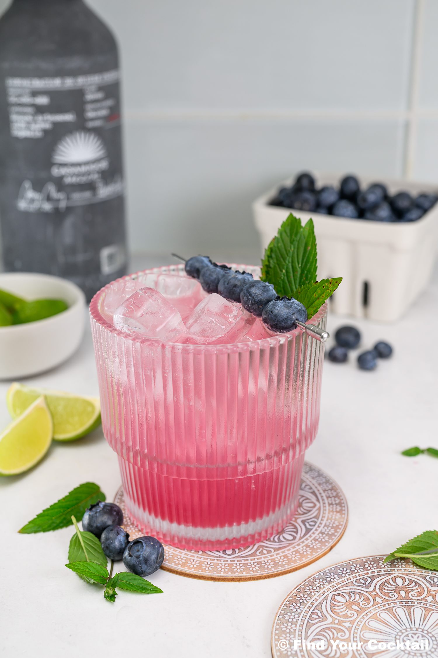 A pink cocktail with ice is garnished with blueberries and mint. Blueberries, lime wedges, and a bottle are in the background on a white surface.