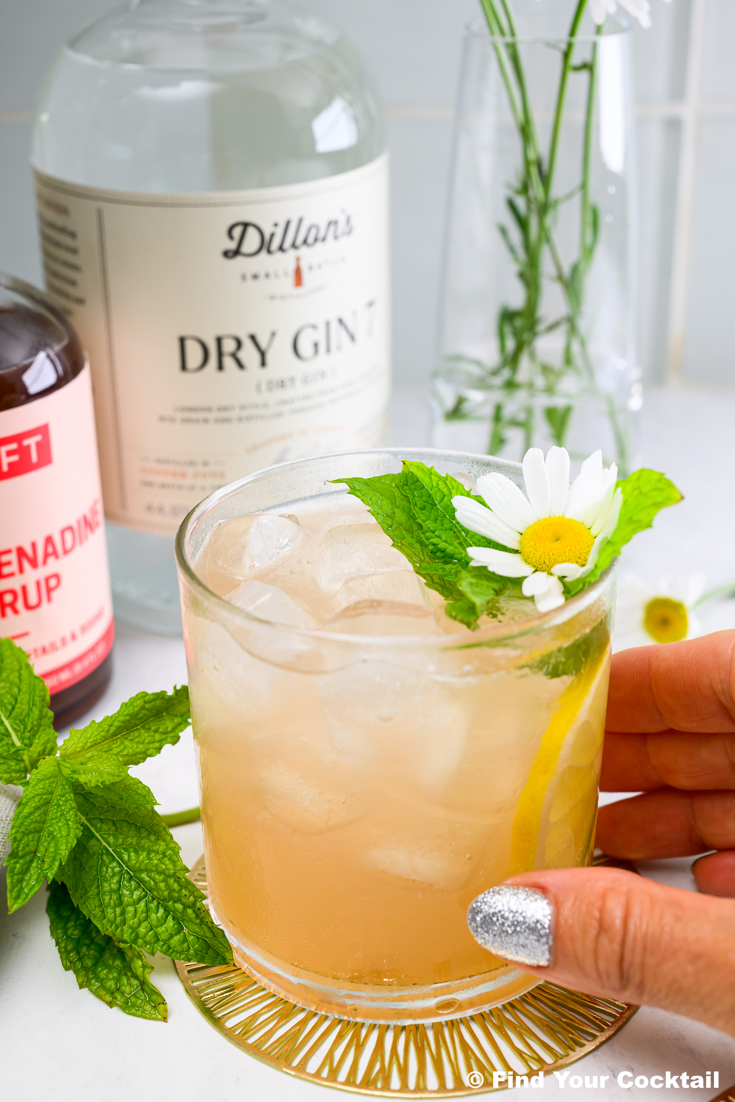 A hand holds a glass of iced cocktail garnished with mint and a daisy, with bottles of gin and grenadine, mint leaves, and a vase of flowers in the background.