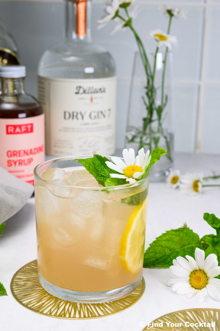A glass of cocktail with ice, lemon slice, mint, and a daisy flower garnish sits on a coaster, with gin, grenadine syrup, and flowers in the background.