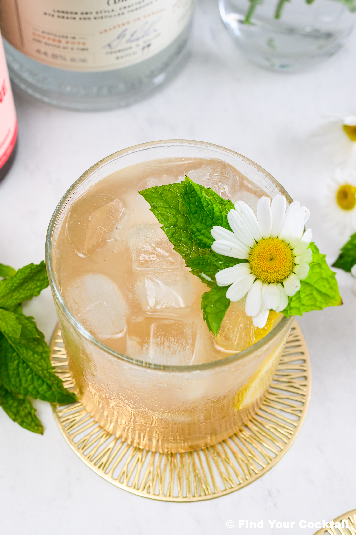 A glass of iced cocktail garnished with a fresh mint sprig and a white daisy, placed on a gold coaster with bottles in the background.