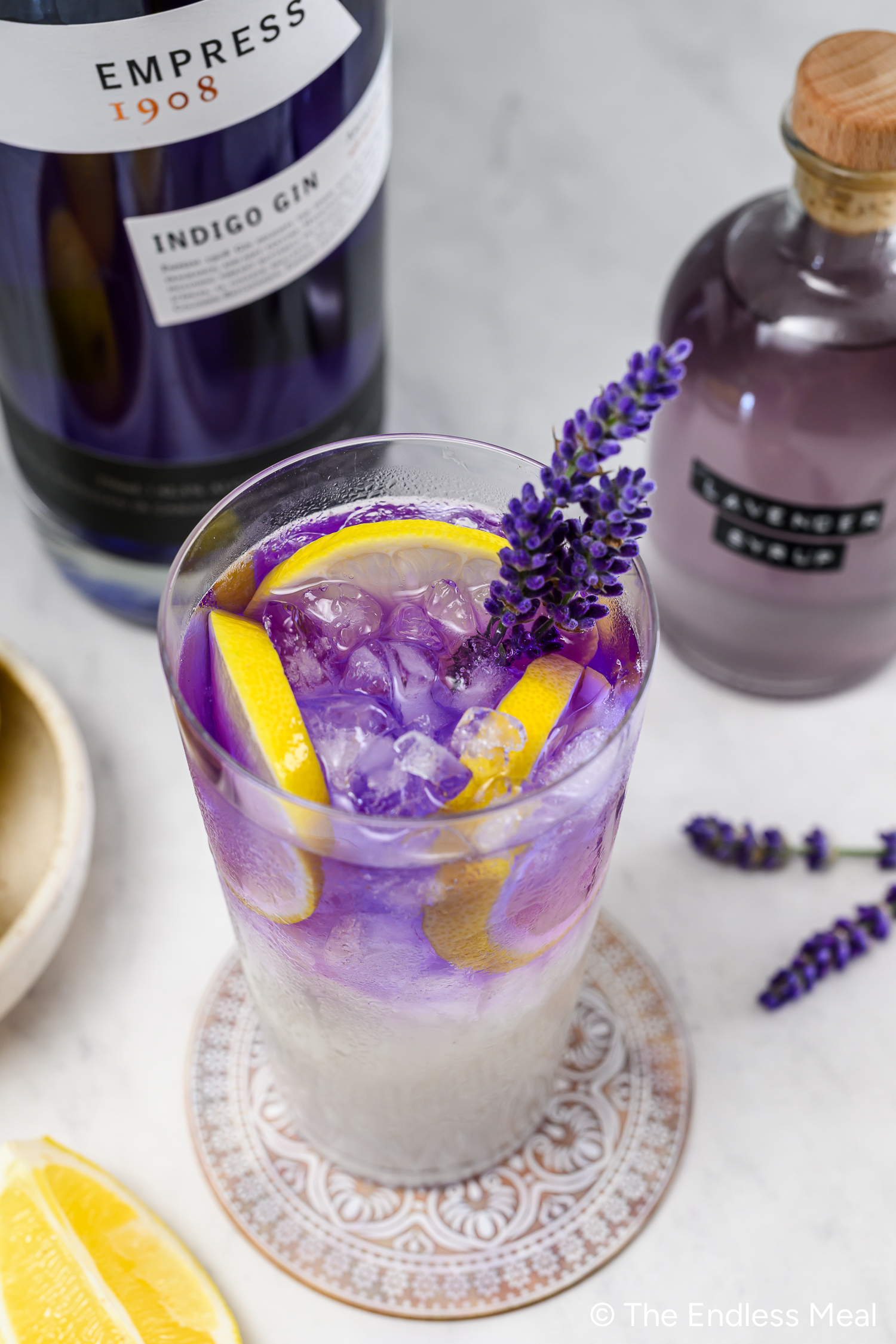 A glass of purple gin cocktail with ice, lemon slices, and a sprig of lavender, next to bottles of Empress 1908 gin and syrup on a white surface.