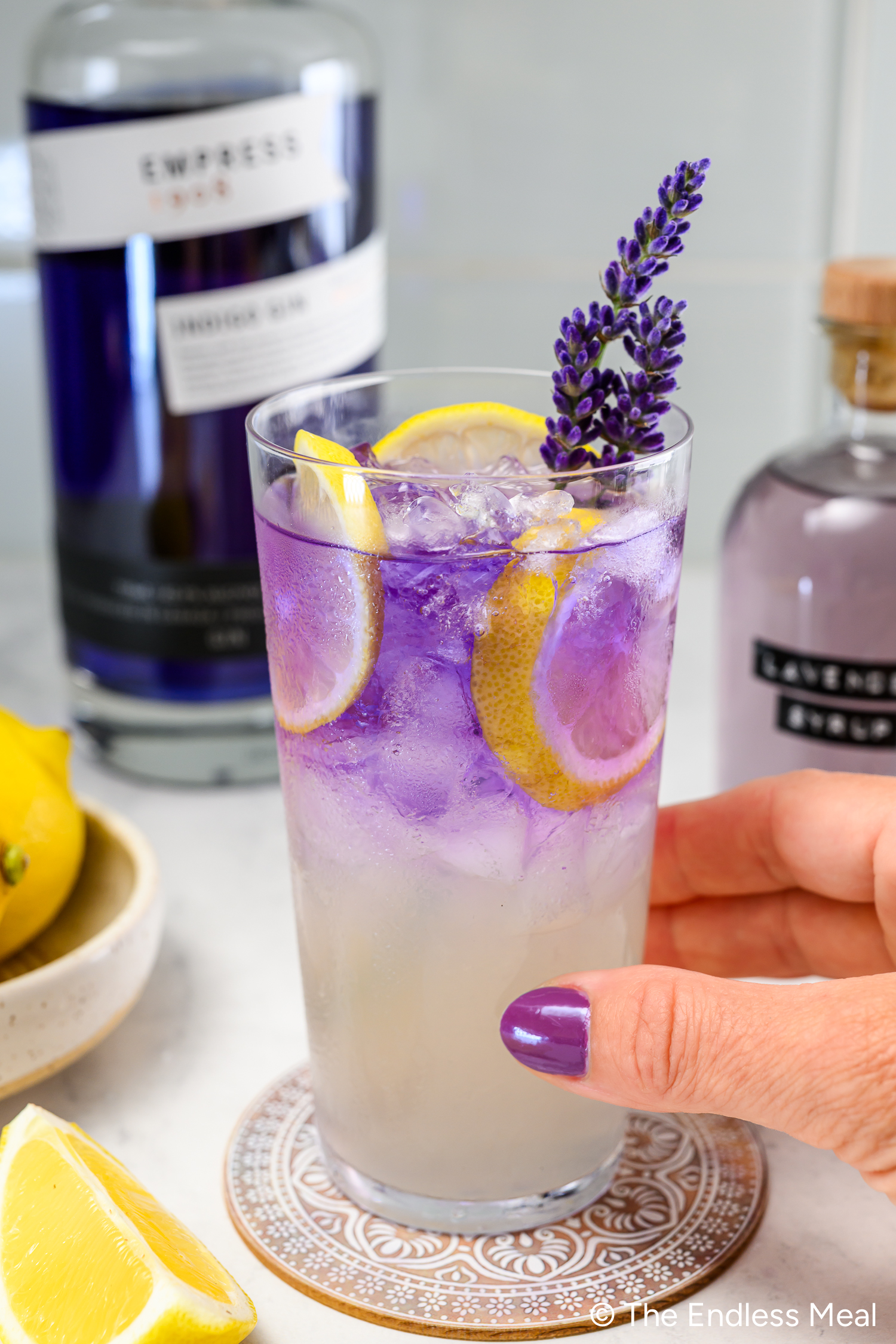 A hand holds a glass of purple-hued cocktail with ice, lemon slices, and a sprig of lavender; bottles and lemons are in the background.