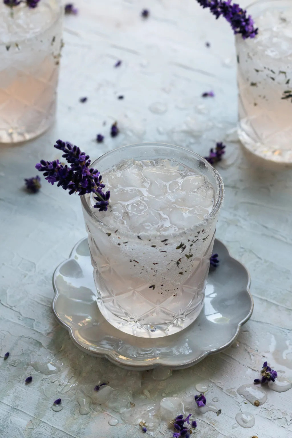 A glass of iced lavender drink garnished with a sprig of lavender, placed on a small white saucer, with scattered lavender flowers on the table.