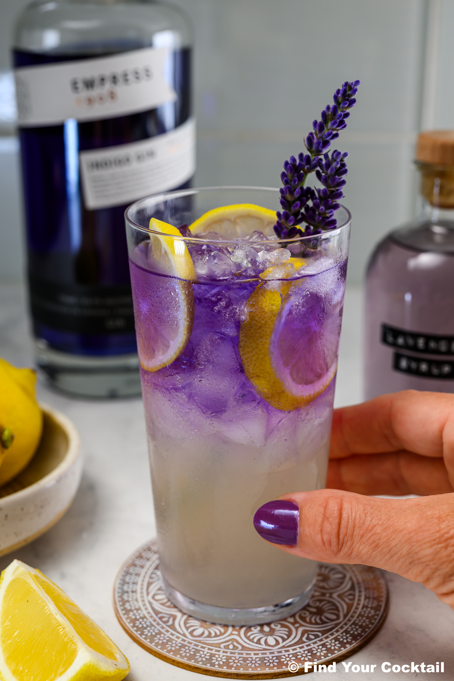 A hand with purple nail polish holds a glass of iced cocktail garnished with lemon slices and a lavender sprig; bottles and lemons are in the background.