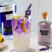 A tall glass of purple and yellow cocktail with lemon slices, ice, and a lavender sprig, placed beside bottles of Empress gin and lavender syrup.