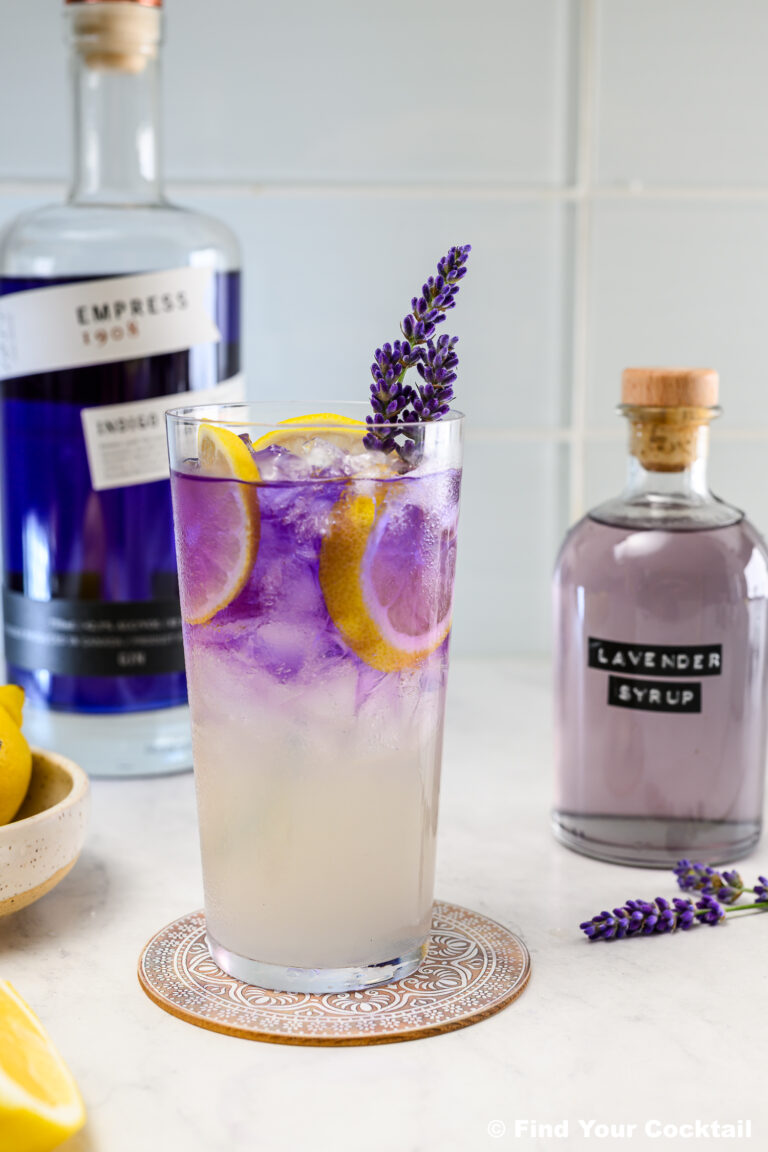 A tall glass of purple and yellow cocktail with lemon slices, ice, and a lavender sprig, placed beside bottles of Empress gin and lavender syrup.