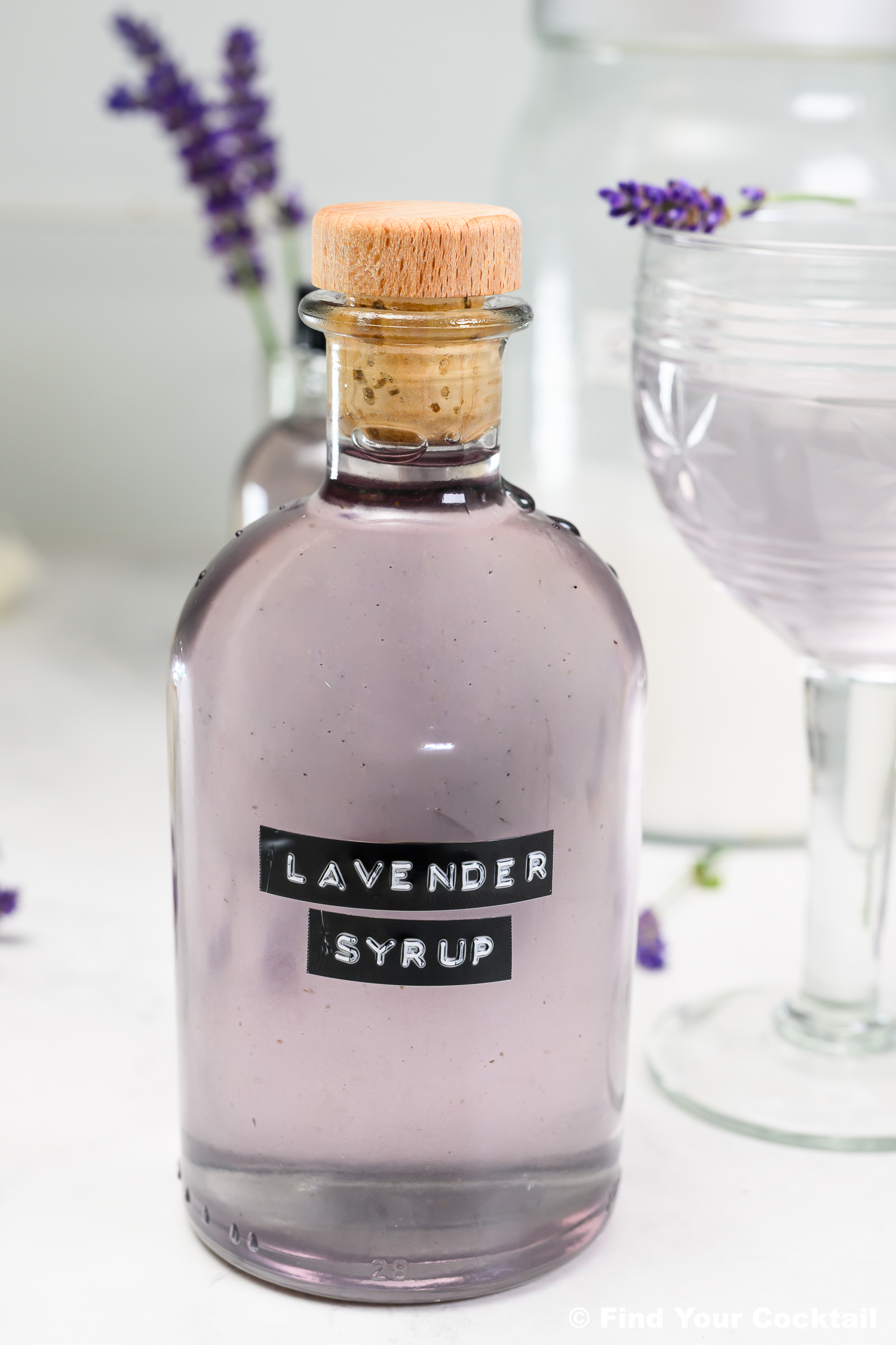 A clear glass bottle labeled Lavender Syrup with a wooden cap sits on a white surface, next to a lavender cocktail and sprigs of fresh lavender.