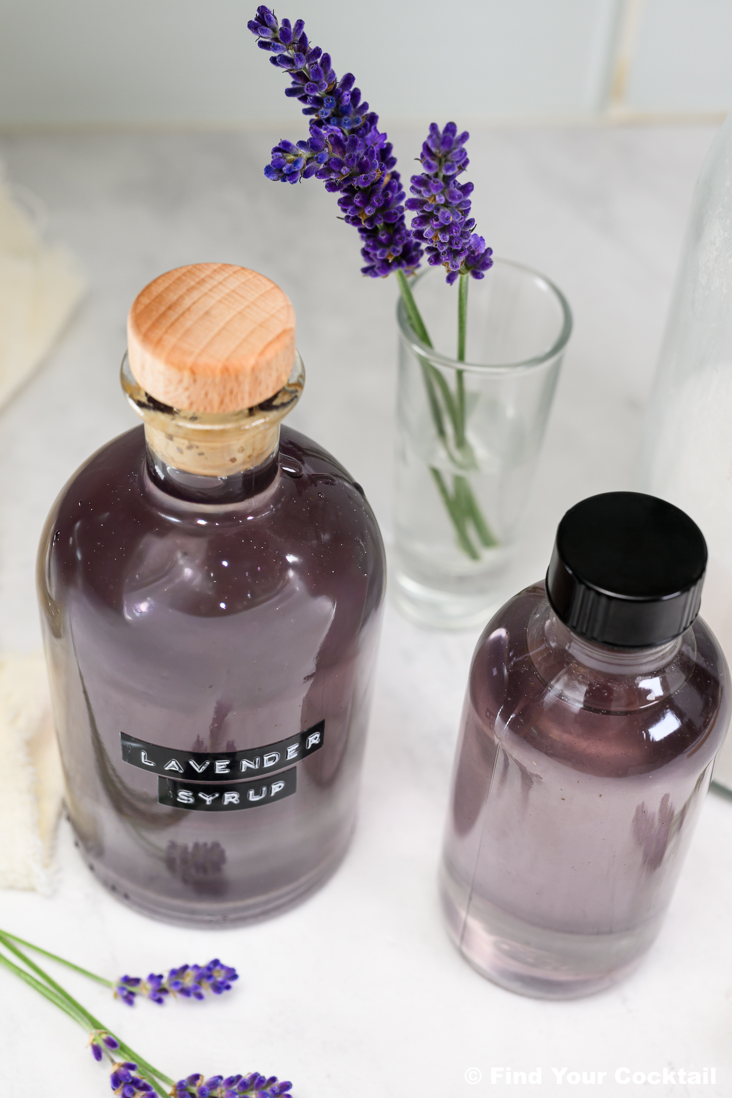 Two bottles of lavender syrup sit on a white surface, one with a cork and wooden cap, next to sprigs of fresh lavender in a glass and on the table.