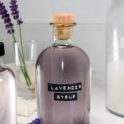 A glass bottle labeled Lavender Syrup with a wooden cork sits on a countertop, surrounded by lavender sprigs and other bottles.