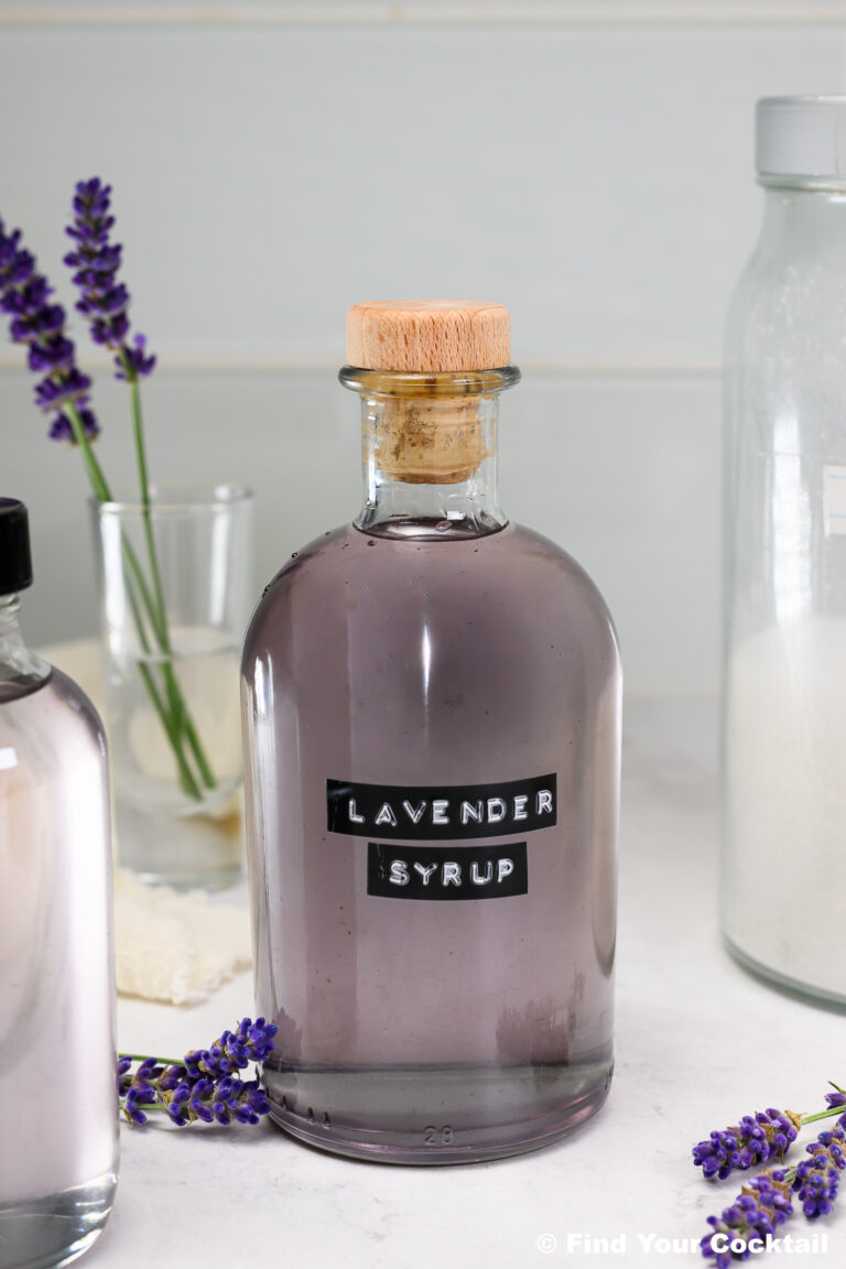 A glass bottle labeled Lavender Syrup with a wooden cork sits on a countertop, surrounded by lavender sprigs and other bottles.