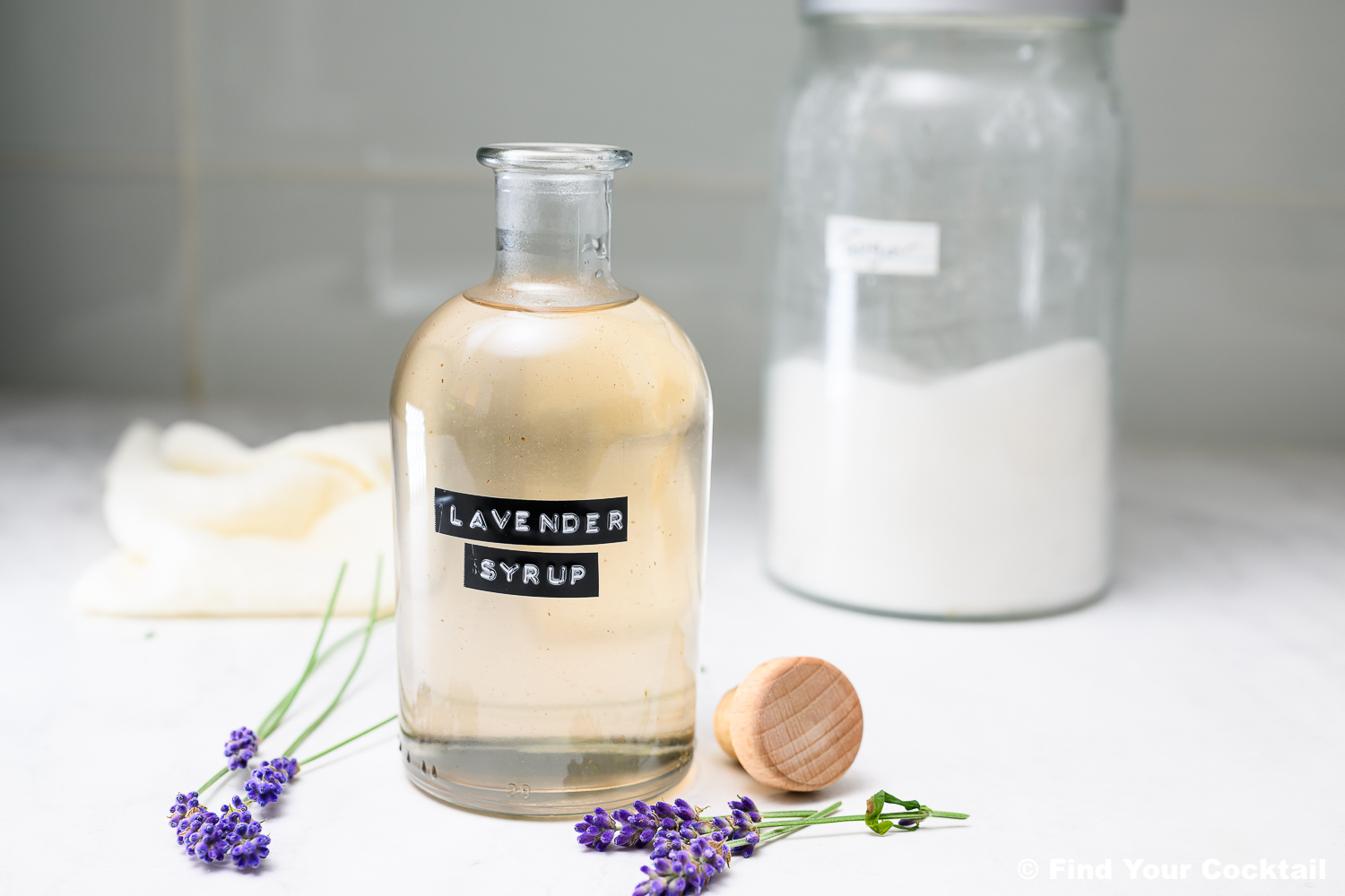 A clear glass bottle labeled Lavender Syrup sits on a counter with a wooden cap nearby, fresh lavender sprigs, a jar of sugar, and a towel in the background.