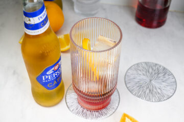 A glass with ice, an orange slice, and a red liquid sits on a coaster next to a bottle of Peroni beer on a marble surface.
