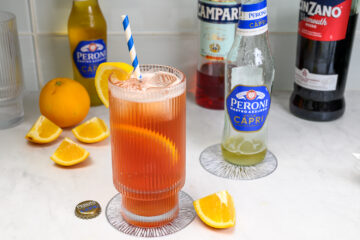 A tall glass of orange-red cocktail with a striped straw and orange slice sits on a coaster, surrounded by orange wedges, bottles, and a Peroni bottle cap on a white surface.
