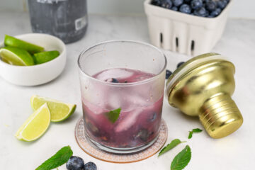 A glass of purple cocktail with ice, blueberries, and mint sits on a coaster next to lime wedges, a gold shaker, loose mint leaves, and a container of blueberries.