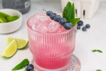 A pink cocktail in a glass with ice, garnished with a metal pick of blueberries and a mint sprig. Lime wedges and loose blueberries are on the white surface nearby.