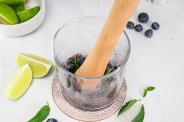 A wooden muddler presses blueberries and mint in a glass, with lime wedges, berries, and mint leaves scattered on a white surface nearby.