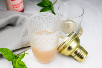 A woven-pattern cocktail shaker with a light pink drink sits on a counter beside its gold lid, a glass, mint leaves, and a gray napkin.