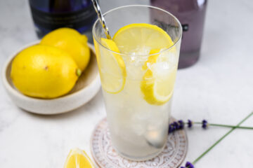 A glass of iced lemonade with lemon slices and a metal stirrer, next to whole lemons and lavender sprigs on a white surface.