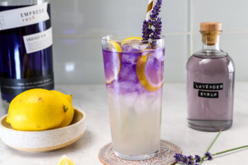 A tall glass of iced lemonade with purple liquid, lemon slices, and a lavender sprig, next to a bottle of lavender syrup, a bowl of lemons, and a bottle of indigo gin.